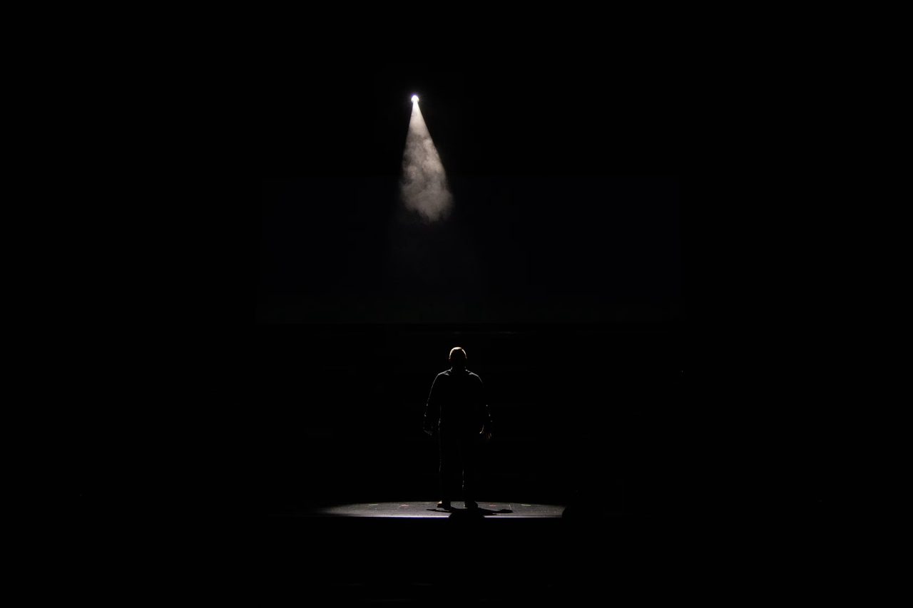 A lone performer stands on a dark stage beneath a single spotlight